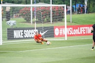 A soccer player wearing a red jersey and orange gloves is crouching on the grass inside the goal area, seemingly in motion to intercept a soccer ball. The goal net and surrounding field are visible in the background. On the left side, part of an advertising board with 'NIKE SOCCER' text is displayed, along with a hashtag referencing a TV network. Spectators and greenery area are visible in the distance.