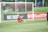 A soccer player wearing a red jersey and orange gloves is crouching on the grass inside the goal area, seemingly in motion to intercept a soccer ball. The goal net and surrounding field are visible in the background. On the left side, part of an advertising board with 'NIKE SOCCER' text is displayed, along with a hashtag referencing a TV network. Spectators and greenery area are visible in the distance.