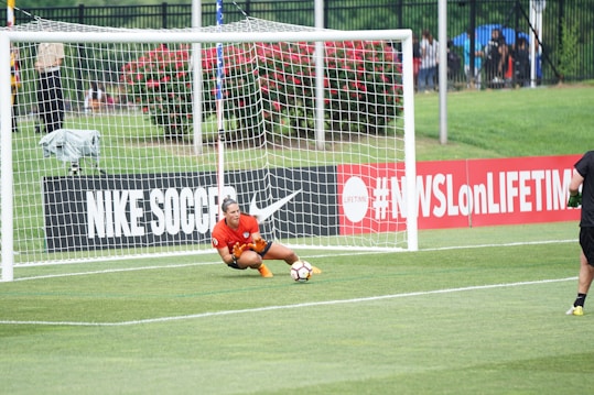 A soccer player wearing a red jersey and orange gloves is crouching on the grass inside the goal area, seemingly in motion to intercept a soccer ball. The goal net and surrounding field are visible in the background. On the left side, part of an advertising board with 'NIKE SOCCER' text is displayed, along with a hashtag referencing a TV network. Spectators and greenery area are visible in the distance.