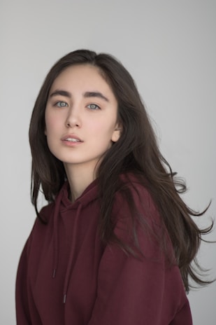A person with long dark hair is wearing a maroon hooded sweatshirt, looking calmly at the camera against a plain, light background.