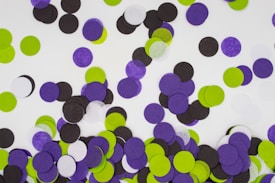 A scattered arrangement of round paper confetti pieces in various colors including black, purple, green, and white placed on a white surface. The confetti pieces are small and circular, creating a playful and festive appearance.