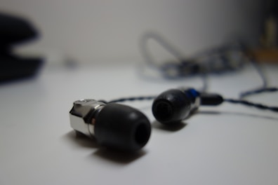 A pair of black and silver in-ear headphones with a braided cable is placed on a light surface. The background is blurred, highlighting the earbuds in the foreground.