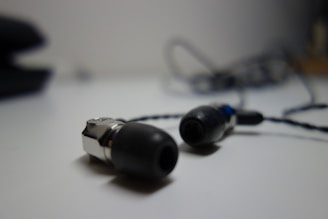 A pair of black and silver in-ear headphones with a braided cable is placed on a light surface. The background is blurred, highlighting the earbuds in the foreground.