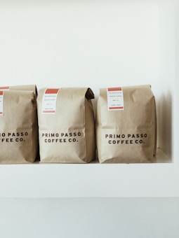 Three brown paper coffee bags with labels sit on a white shelf. The bags are from Primo Passo Coffee Co.
