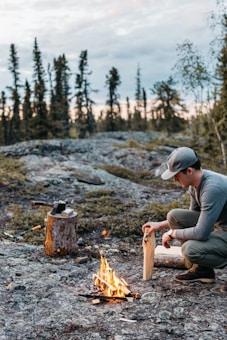 A person is crouching and preparing firewood beside a small campfire on a rocky terrain. In the background, there are tall, sparse trees and a slightly cloudy sky, suggesting an outdoor camping scene. An axe is lodged into a tree stump nearby.