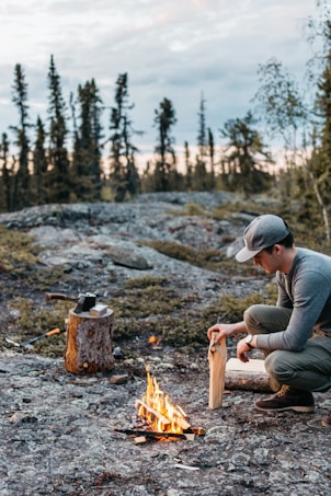 A person is crouching and preparing firewood beside a small campfire on a rocky terrain. In the background, there are tall, sparse trees and a slightly cloudy sky, suggesting an outdoor camping scene. An axe is lodged into a tree stump nearby.