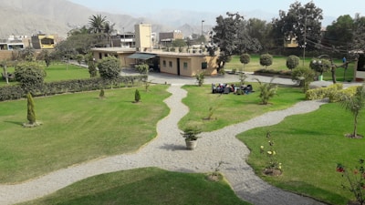 A well-maintained garden with lush green grass, several pathways converging and diverging, bordered by small shrubs and neatly trimmed trees. In the background, a small beige building is partially visible, surrounded by scattered trees. There is a group of people sitting or lying on the grass near the center of the garden.