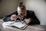 A person with a topknot hairstyle intensely examines a book using a magnifying glass. Their attire includes a dark, long-sleeved garment, and they wear several rings, one of which features a cross. The setting is minimalistic, with the person leaning over a wooden table in front of a light-colored wall.