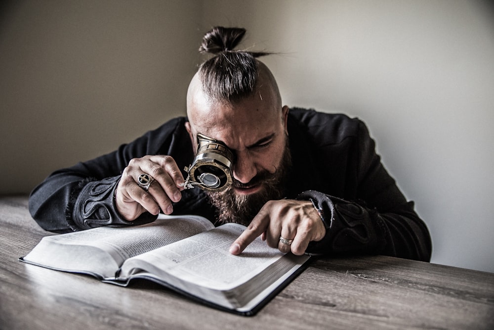 man holding reading magnifying glass rearing a book on brown wooden table