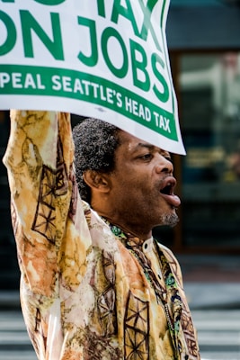 A person wearing a patterned shirt is actively participating in a protest or demonstration, holding up a sign with text related to taxes and jobs.