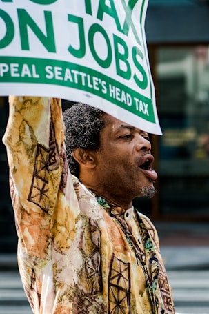 A person wearing a patterned shirt is actively participating in a protest or demonstration, holding up a sign with text related to taxes and jobs.
