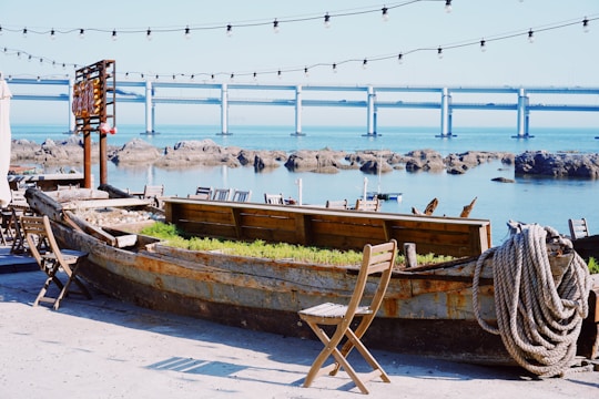 An old wooden boat repurposed as a planter with green foliage is situated near the waterfront. Rustic wooden chairs and tables are arranged around it, creating a cozy seating area. Overhead string lights add a warm ambiance. Beyond the patio, large rocks and a peaceful body of water can be seen, along with a modern bridge stretching across the horizon.