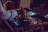A person operates a DJ controller, adjusting the knobs and sliders on the equipment. The setup is illuminated by small, colorful LED lights, creating a vibrant and dynamic atmosphere.