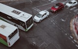 Aerial view of an urban road intersection featuring a large white and green bus, a line of three cars including a white sedan, white compact car, and a red SUV. The scene includes painted road markings and a nearby red bus stop.