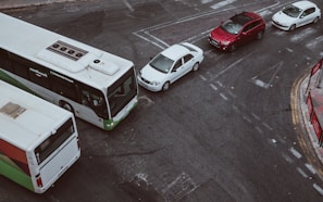 Aerial view of an urban road intersection featuring a large white and green bus, a line of three cars including a white sedan, white compact car, and a red SUV. The scene includes painted road markings and a nearby red bus stop.