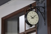 An antique-style wall clock with Roman numerals is mounted on a bracket attached to a wall, adjacent to a window with a wooden frame. The clock has a vintage appearance with decorative hands.