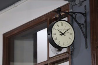 An antique-style wall clock with Roman numerals is mounted on a bracket attached to a wall, adjacent to a window with a wooden frame. The clock has a vintage appearance with decorative hands.
