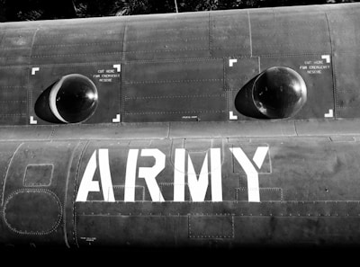 A portion of a military aircraft is visible with the word 'ARMY' prominently displayed in bold white letters on a dark surface. There are two circular, dome-like structures with markings indicating emergency cut points. The surface has detailed panel lines and rivets, contributing to an industrial and military aesthetic.
