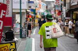 A sunlit scene showing a woman carrying a vivid tote bag while walking through a lively street.