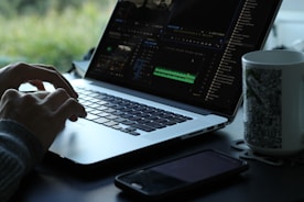 A person is using a laptop with a video editing software open on the screen. The workspace is minimalistic, featuring a smartphone and a mug with a black and white pattern. Natural light filters in, creating a serene atmosphere with greenery visible in the background.