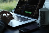 A person is using a laptop with a video editing software open on the screen. The workspace is minimalistic, featuring a smartphone and a mug with a black and white pattern. Natural light filters in, creating a serene atmosphere with greenery visible in the background.