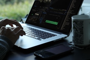 A person is using a laptop with a video editing software open on the screen. The workspace is minimalistic, featuring a smartphone and a mug with a black and white pattern. Natural light filters in, creating a serene atmosphere with greenery visible in the background.