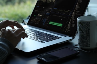 A person is using a laptop with a video editing software open on the screen. The workspace is minimalistic, featuring a smartphone and a mug with a black and white pattern. Natural light filters in, creating a serene atmosphere with greenery visible in the background.