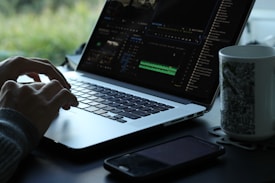 A person is using a laptop with a video editing software open on the screen. The workspace is minimalistic, featuring a smartphone and a mug with a black and white pattern. Natural light filters in, creating a serene atmosphere with greenery visible in the background.