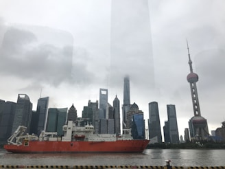 A large red cargo ship sails through a river with a backdrop of modern skyscrapers. The skyline includes various uniquely shaped buildings under a cloudy sky, with the prominent Oriental Pearl Tower visible on the right.