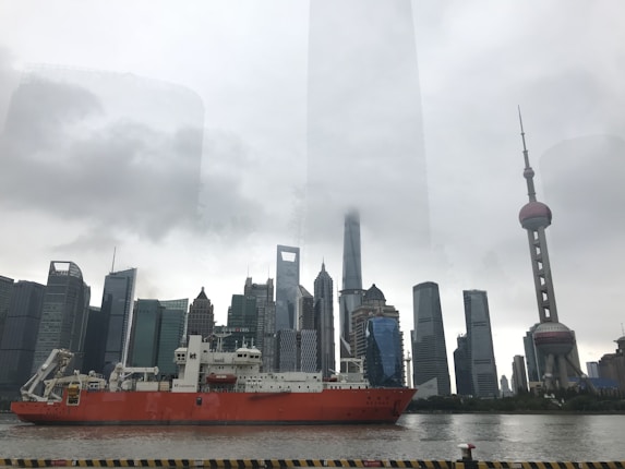 A large red cargo ship sails through a river with a backdrop of modern skyscrapers. The skyline includes various uniquely shaped buildings under a cloudy sky, with the prominent Oriental Pearl Tower visible on the right.