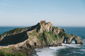 A rocky island landscape featuring a winding stone pathway leading up to a small building perched atop the island. The island is surrounded by the ocean, with waves crashing against its base. Green foliage covers parts of the rocky surface, adding natural color to the scene.