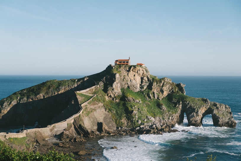 A rocky island landscape featuring a winding stone pathway leading up to a small building perched atop the island. The island is surrounded by the ocean, with waves crashing against its base. Green foliage covers parts of the rocky surface, adding natural color to the scene.