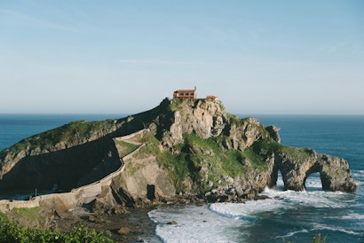 A rocky island landscape featuring a winding stone pathway leading up to a small building perched atop the island. The island is surrounded by the ocean, with waves crashing against its base. Green foliage covers parts of the rocky surface, adding natural color to the scene.