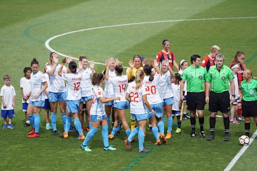 A group of female soccer players in white and blue uniforms are gathered on a green soccer field. They appear to be celebrating or strategizing before a match. Several referees in green shirts stand nearby while ball boys and girls in white t-shirts and blue shorts are present as well.