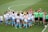 A group of female soccer players in white and blue uniforms are gathered on a green soccer field. They appear to be celebrating or strategizing before a match. Several referees in green shirts stand nearby while ball boys and girls in white t-shirts and blue shorts are present as well.