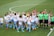 A group of female soccer players in white and blue uniforms are gathered on a green soccer field. They appear to be celebrating or strategizing before a match. Several referees in green shirts stand nearby while ball boys and girls in white t-shirts and blue shorts are present as well.