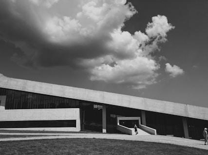 A modern architectural building with angular lines stands beneath a dramatic sky filled with large, fluffy clouds. The structure has a sleek, concrete facade and expansive glass windows. People are walking on the pathway leading to the building, which is surrounded by a grassy area.