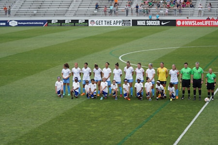 A group of soccer players and officials are standing and kneeling on a large soccer field. The players are wearing white jerseys and blue shorts, while the officials are in green. The background shows empty bleachers with a few spectators scattered around.