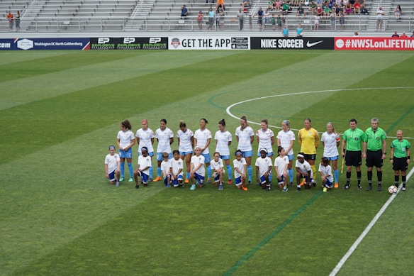 A group of soccer players and officials are standing and kneeling on a large soccer field. The players are wearing white jerseys and blue shorts, while the officials are in green. The background shows empty bleachers with a few spectators scattered around.