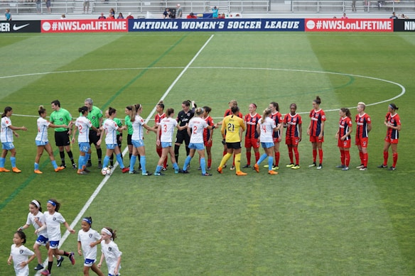 Two women's soccer teams, one in white and blue uniforms and the other in red and black uniforms, are shaking hands on a green soccer field. Referees wearing green shirts are also present. Young children wearing white outfits are running in the foreground, and a banner promoting a women's soccer league is visible in the background.