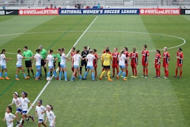 Two women's soccer teams, one in white and blue uniforms and the other in red and black uniforms, are shaking hands on a green soccer field. Referees wearing green shirts are also present. Young children wearing white outfits are running in the foreground, and a banner promoting a women's soccer league is visible in the background.