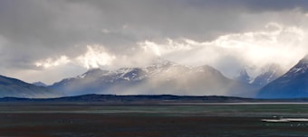 The image features a dramatic landscape with snow-capped mountains partially obscured by mist and clouds. The foreground shows a flat terrain with patches of green and brown earth, hinting at a sparse, possibly tundra-like environment. The clouds are thick and varied, creating dynamic lighting and casting shadows over the mountains.