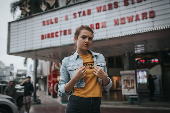 A young person stands confidently in front of a theater marquee that advertises a film by Ron Howard. The individual wears a denim jacket over a yellow graphic tee, with the background showing blurred figures and urban surroundings.