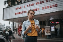 A young person stands confidently in front of a theater marquee that advertises a film by Ron Howard. The individual wears a denim jacket over a yellow graphic tee, with the background showing blurred figures and urban surroundings.
