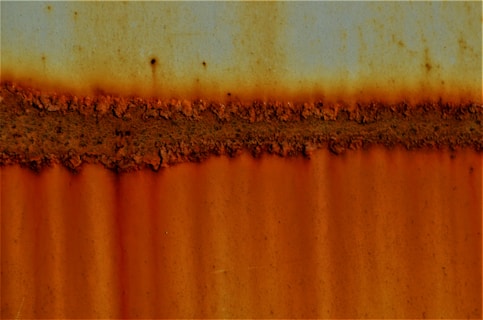 A textured, rust-covered surface with a horizontal band of deep orange corrosion. The image features a blend of muted blue hues at the top, transitioning to varying shades of orange and brown, signifying extensive rust on a metallic surface.