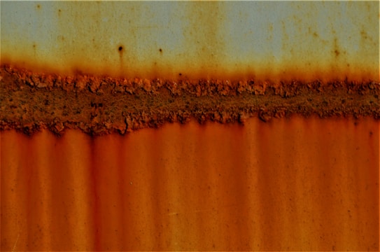A textured, rust-covered surface with a horizontal band of deep orange corrosion. The image features a blend of muted blue hues at the top, transitioning to varying shades of orange and brown, signifying extensive rust on a metallic surface.