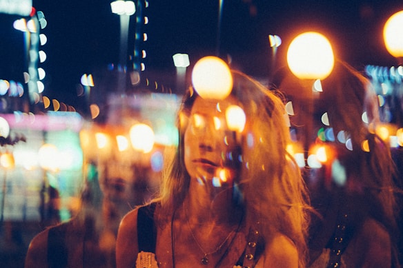 A woman appears in a dreamy, abstracted scene with overlapping bright orbs of light, likely from street lamps or city lights, creating a surreal and artsy effect. The environment suggests a lively urban night setting with a blurred background.