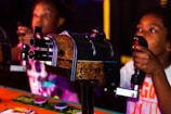 Two young individuals are engaged in an interactive arcade game, holding onto control handles with focus and excitement. The game setup includes a wooden and metal design, creating a lively and immersive atmosphere.