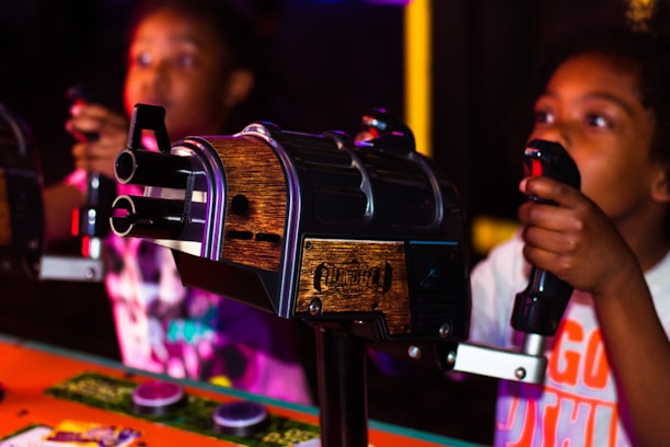 Two young individuals are engaged in an interactive arcade game, holding onto control handles with focus and excitement. The game setup includes a wooden and metal design, creating a lively and immersive atmosphere.