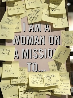 A central poster with the text 'I AM A WOMAN ON A MISSION TO...' is surrounded by numerous sticky notes. Each note contains handwritten motivational messages or goals such as 'Support the Arts!', 'Love and be loved', 'Create beauty!', and 'Make people feel good'. The sticky notes are of various colors, mostly shades of yellow, and are adhered in a seemingly random pattern around the central poster.